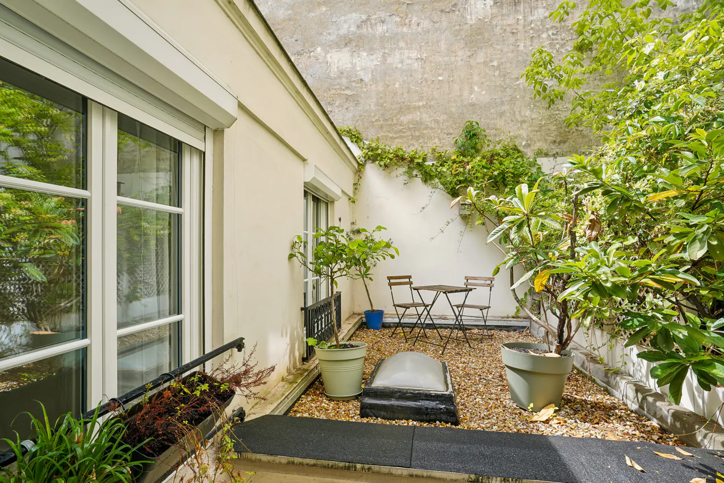 A small outdoor courtyard with a table and chairs, potted plants, and a gravel floor. The courtyard is surrounded by white walls and greenery.