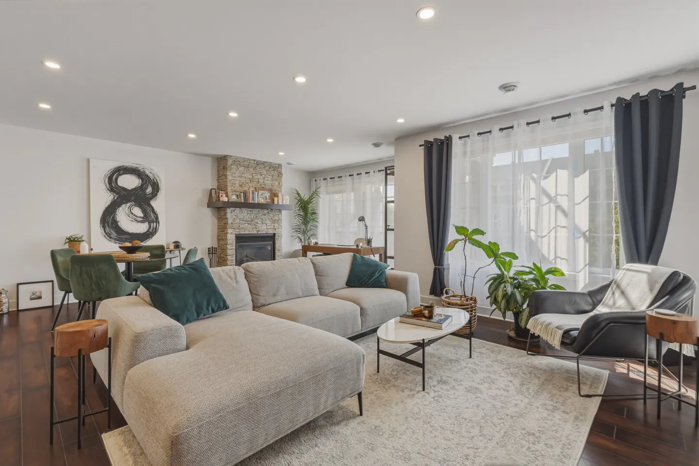 Bright living room with a stone fireplace, beige sectional sofa, and dark wood floors. A black and white painting hangs above a dining table.