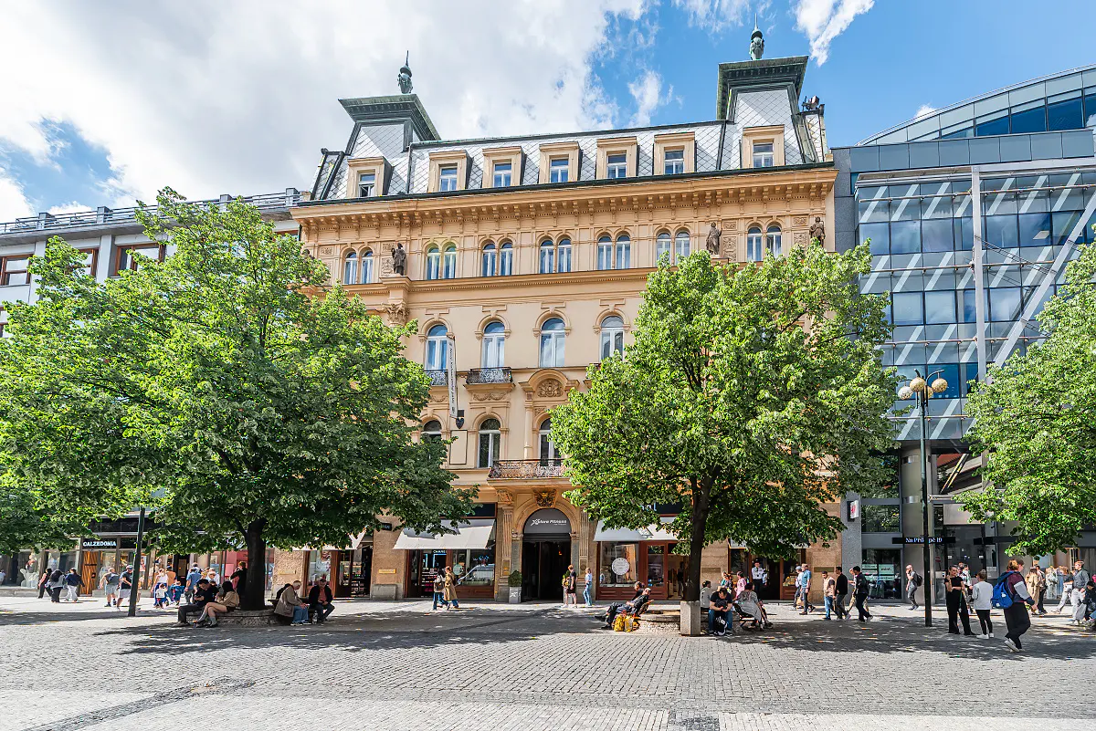 A tan building with a gray roof stands in a plaza with trees and people.