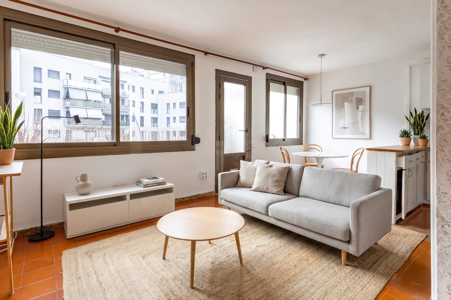 Living room with a gray sofa, round wood table, and jute rug. Windows overlook a city building. Neutral tones and natural light fill the space.