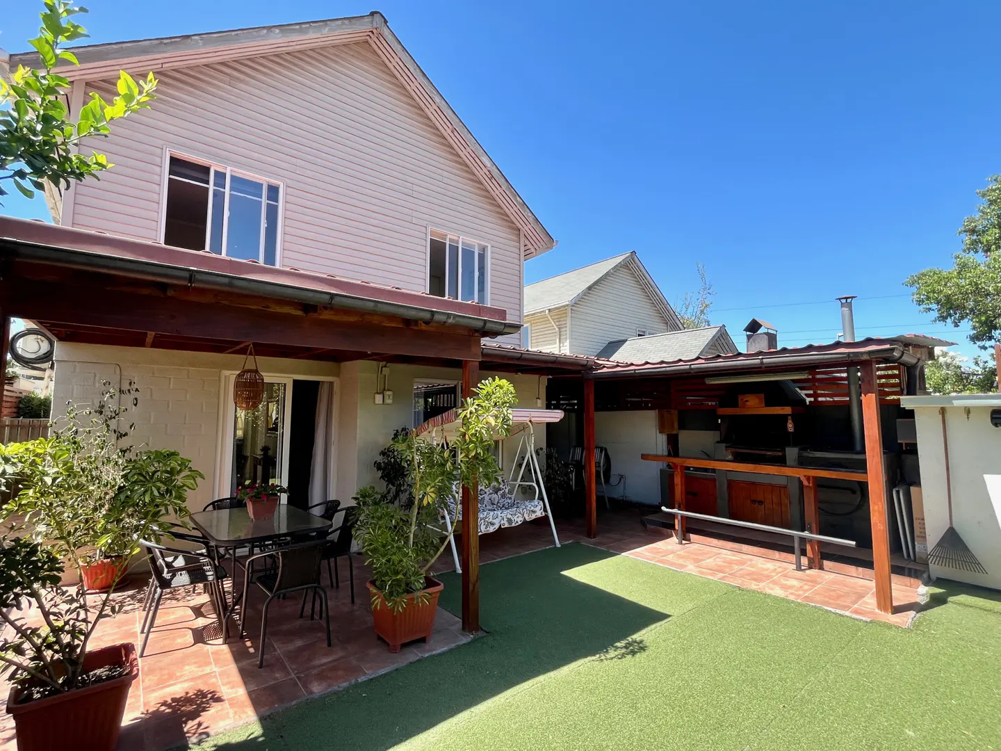Backyard patio with outdoor kitchen, dining table, swing, and artificial grass. The house is two stories with light pink siding.