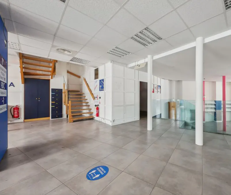 A spacious commercial space with gray tile flooring, white walls, and a wooden staircase. Blue cabinets and a fire extinguisher are visible.