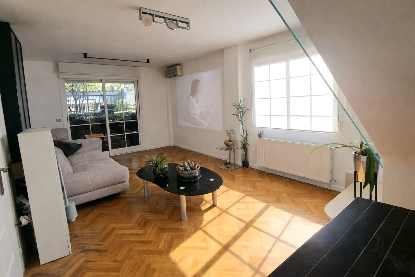 Bright living room with herringbone wood floors, a gray sofa, and a black coffee table. A projector displays an image on the wall. Large windows let in sunlight.