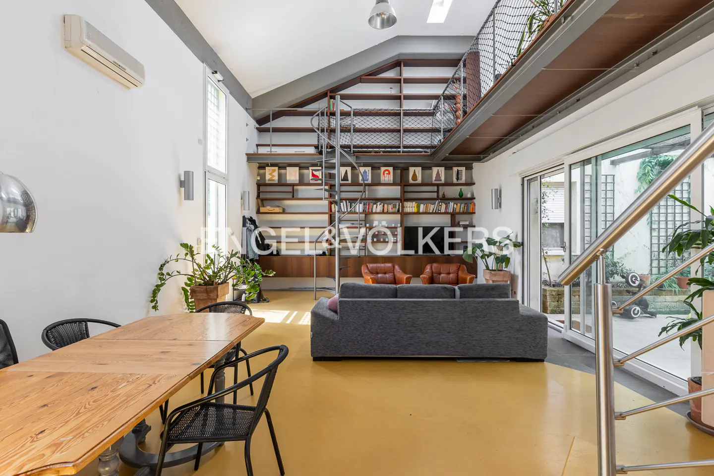 Bright, modern living room with yellow floor, gray sofa, wood table, and spiral staircase leading to a loft with bookshelves. Sliding glass doors to patio.