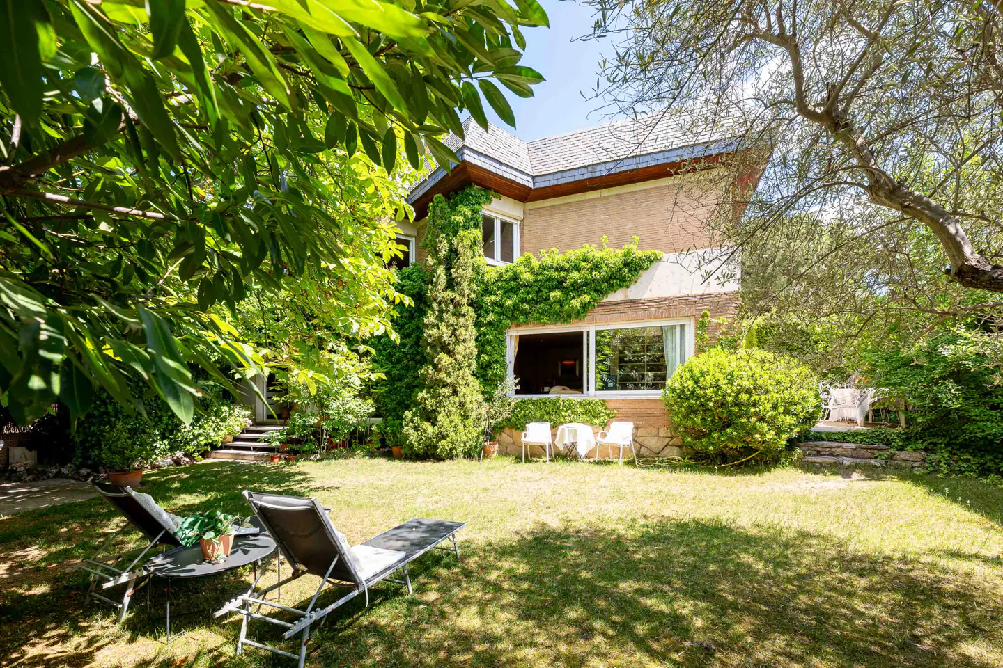 A two-story tan house with green ivy sits behind a lush green lawn with lounge chairs and trees.