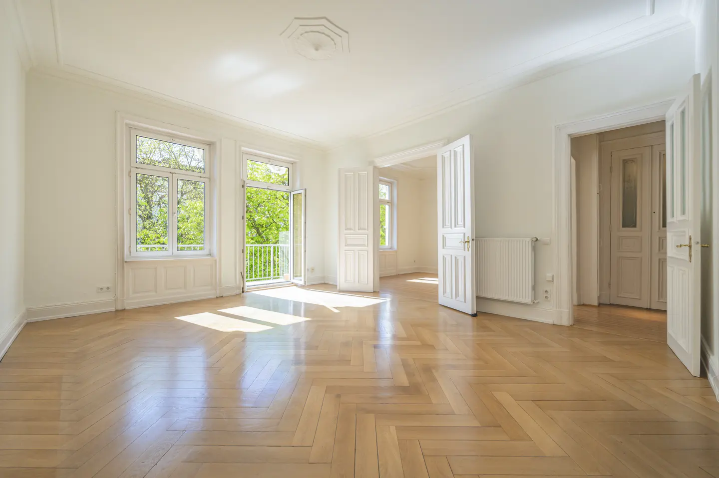 Bright, empty room with white walls, herringbone wood floors, large windows, and an open balcony door overlooking trees.