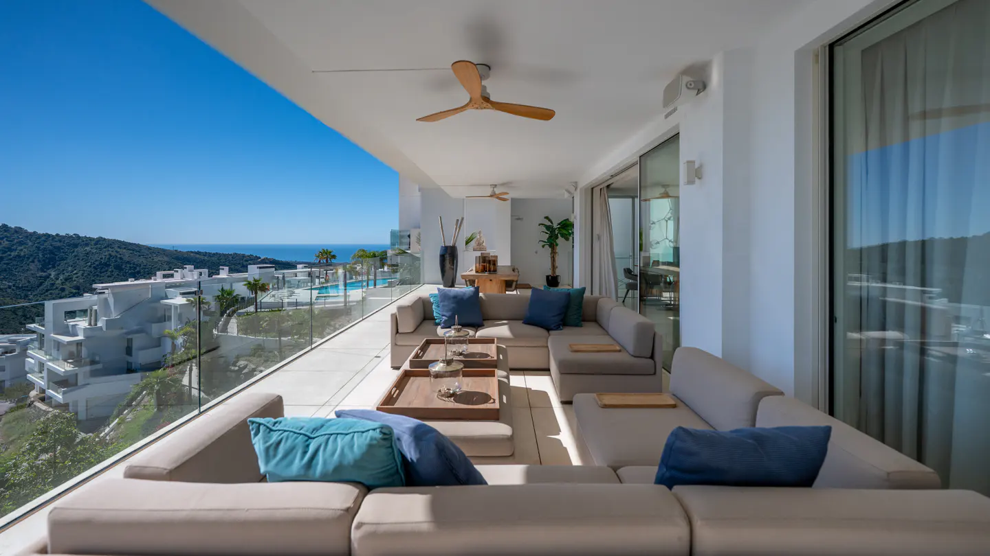 Luxury balcony with beige sectional sofa, blue pillows, and ocean view. Glass railings and a wooden ceiling fan add to the upscale ambiance.