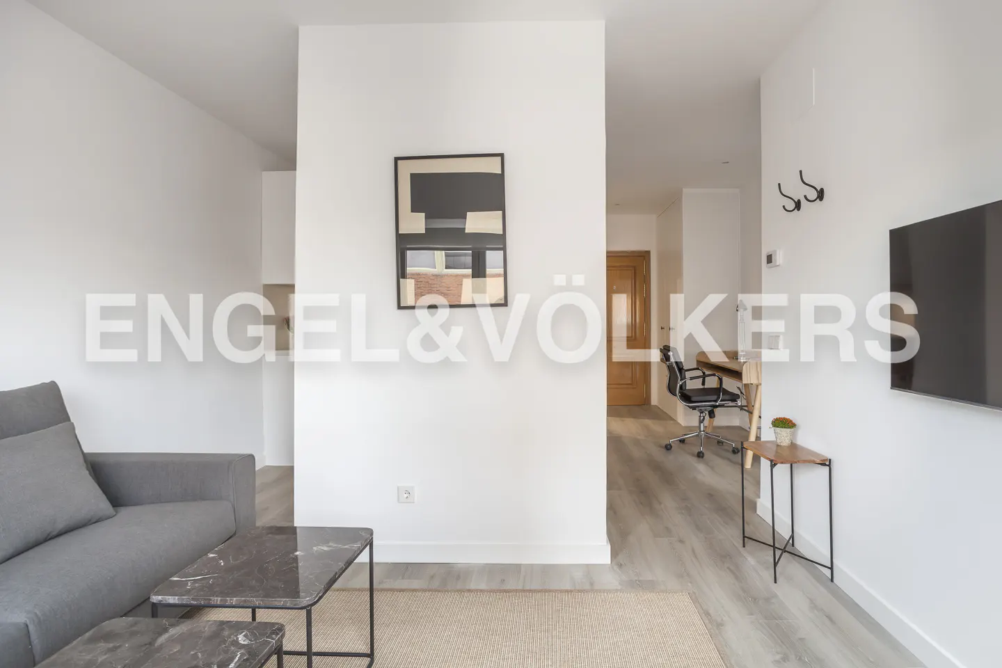 Living room with gray sofa, marble tables, and abstract art on white walls. Desk and chair visible in the background.