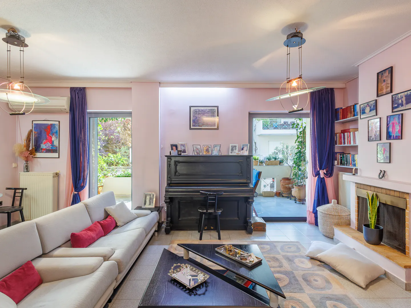 Living room with pink walls, a black piano, and a light gray sofa with red pillows. There is a fireplace, a bookshelf, and a door to a patio.
