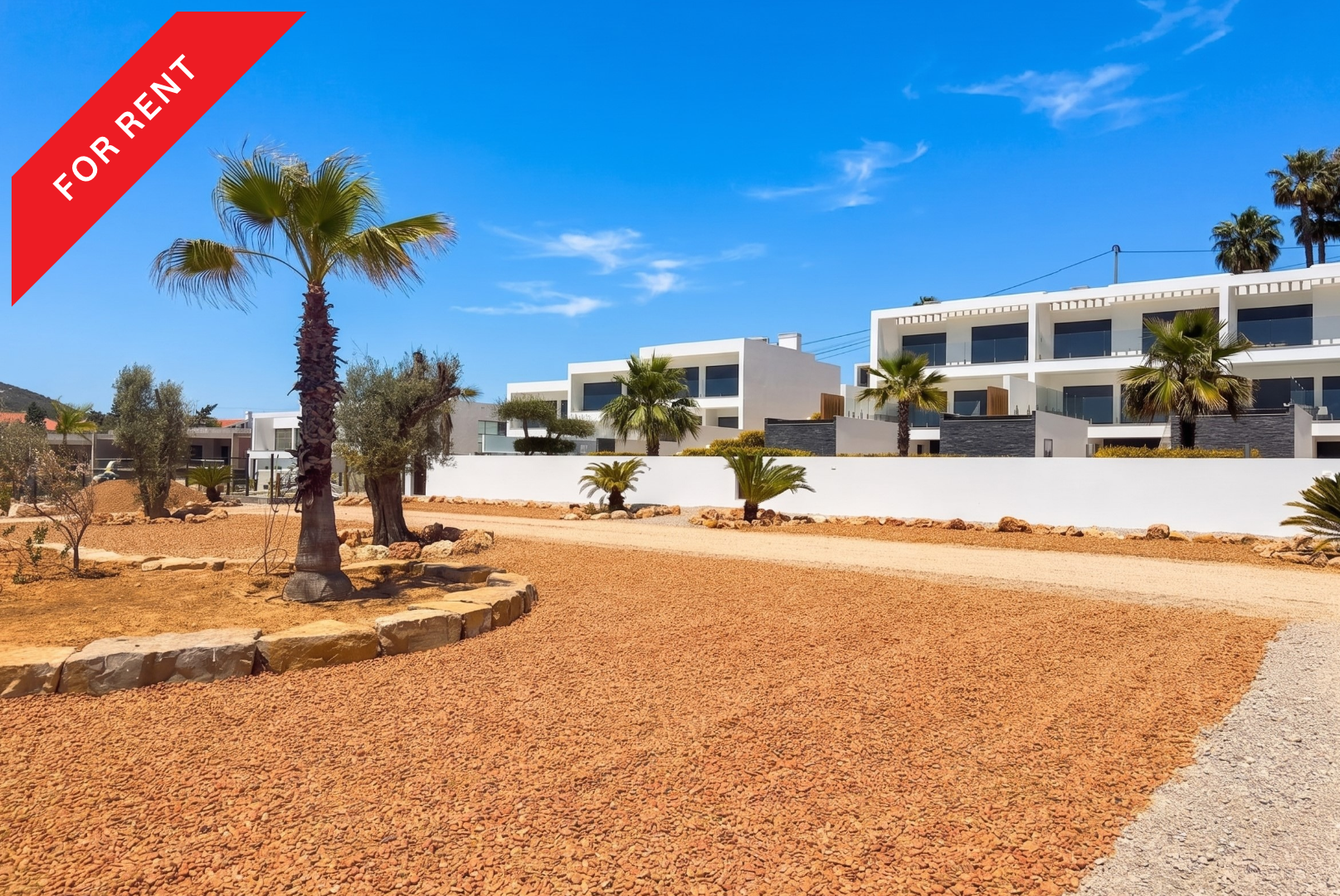 Modern white apartment buildings with palm trees and a gravel yard under a clear blue sky, marked with a For Rent banner. [AI-generated text]