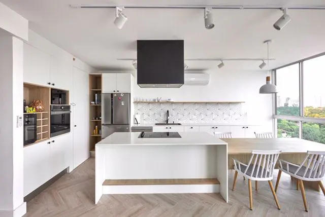Modern white kitchen with a large island, black range hood, herringbone wood floors, and a dining area by large windows.