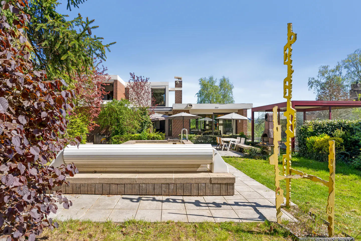 Backyard view of a modern brick home with a covered pool, patio furniture, and a tall yellow sculpture on the green lawn.