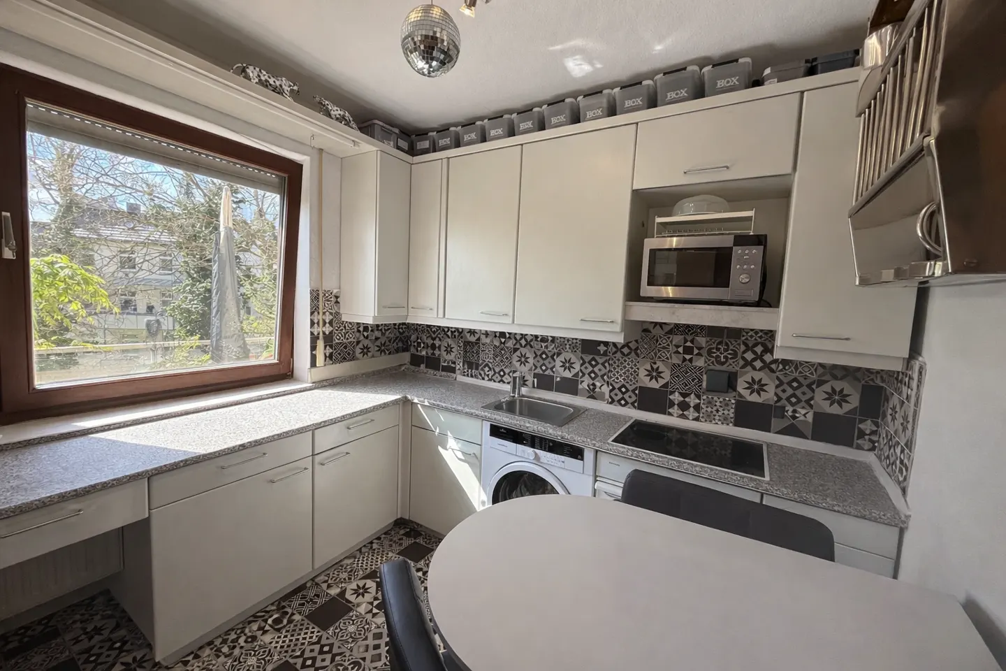 Bright kitchen with white cabinets, patterned backsplash and floor, and a large window. A round white table sits in the foreground.