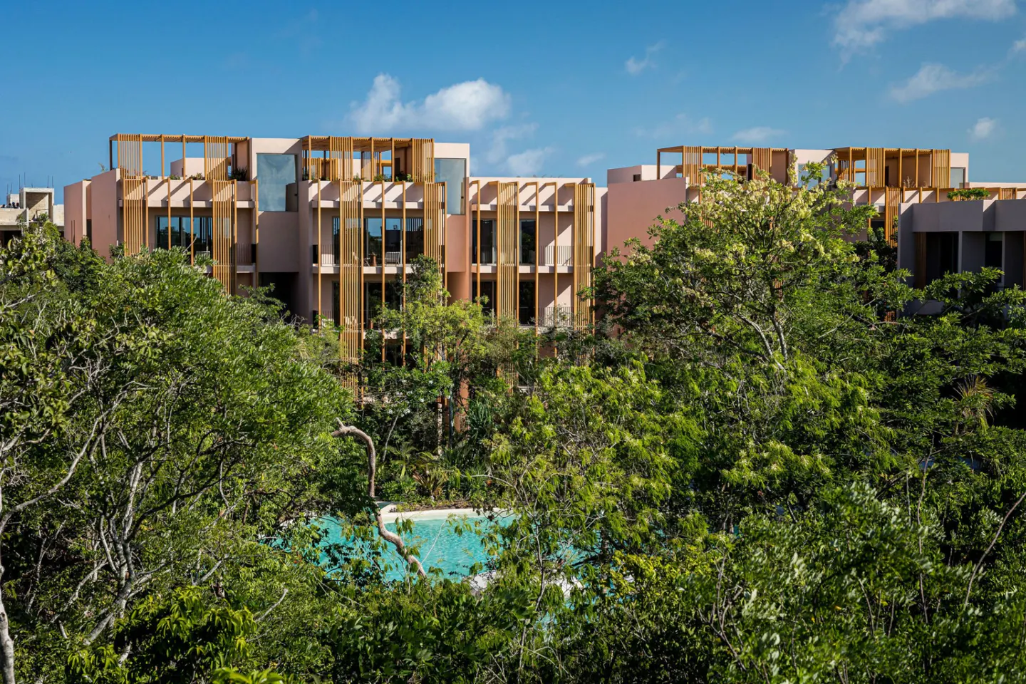 Exterior view of a pink building with wood accents, partially obscured by green trees. A turquoise pool is visible through the trees. Blue sky.
