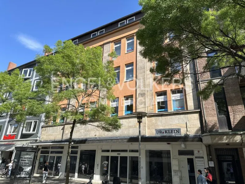 Exterior view of a multi-story building with shops on the ground floor. The building is peach and gray, with trees in front. "Dr. Klein" sign visible.