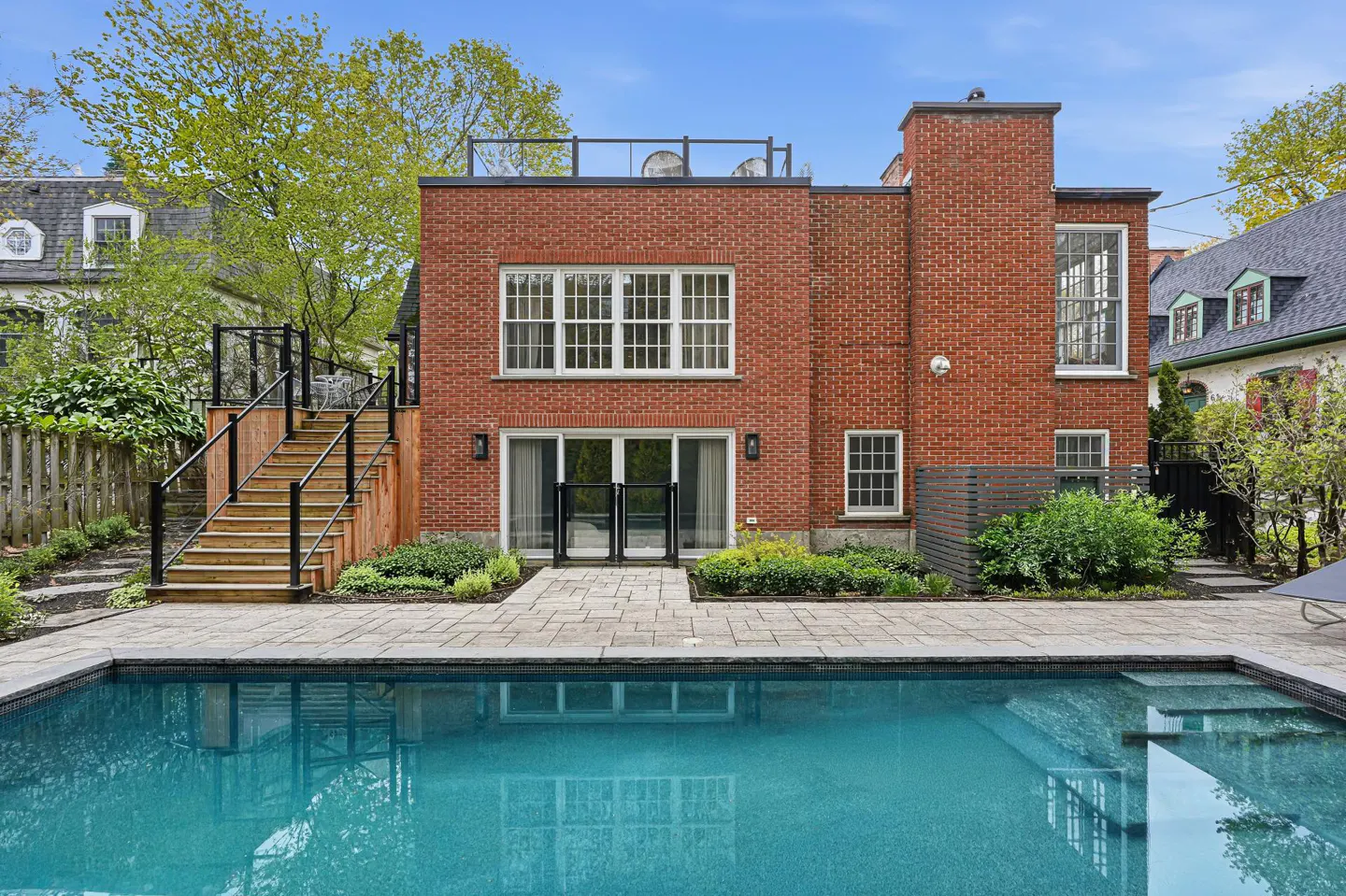 Backyard view of a red brick house with a blue swimming pool in the foreground. A wooden staircase leads to a rooftop deck.