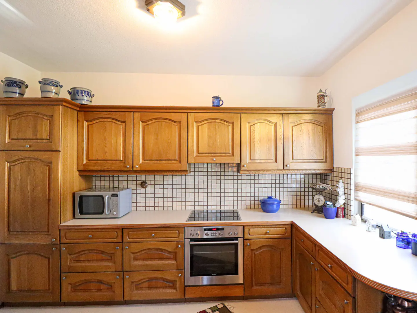 A bright kitchen with light wood cabinets, white countertops, and a black and white tiled backsplash. A microwave and oven are visible.