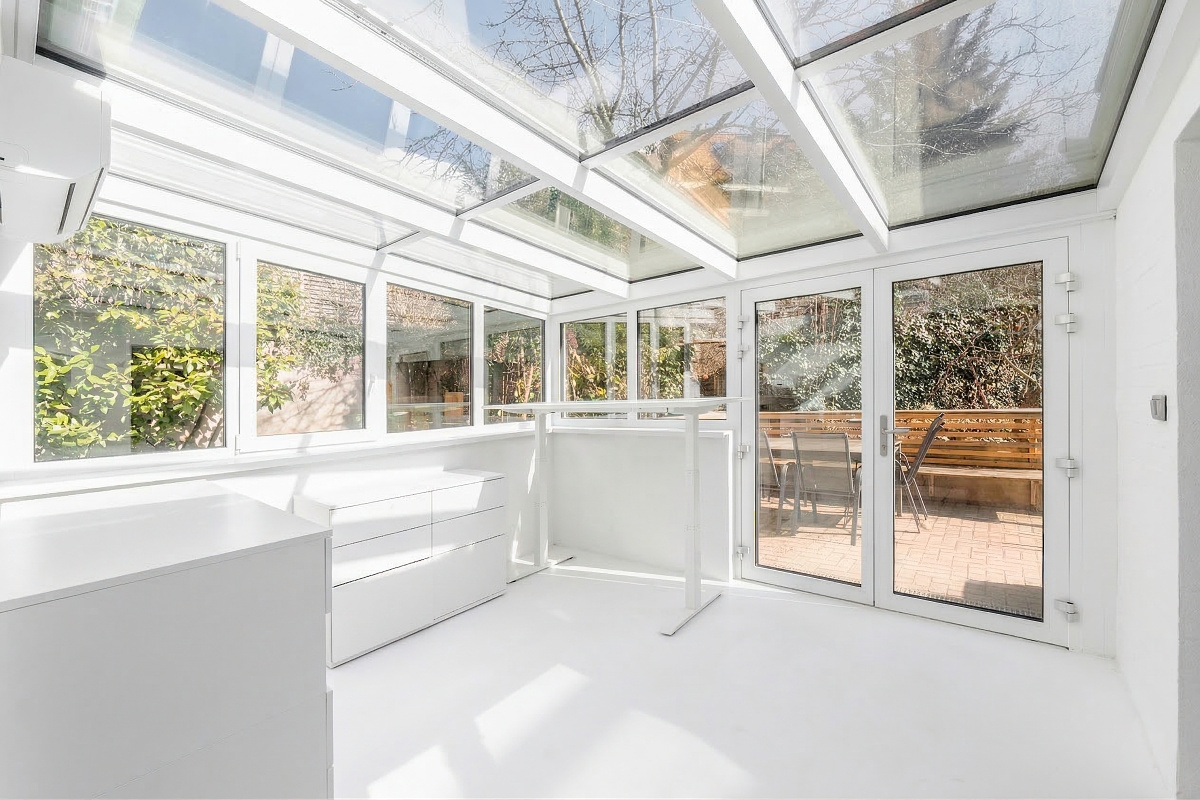 Bright sunroom with white walls, glass ceiling, standing desk, and double doors opening to a patio with outdoor seating.