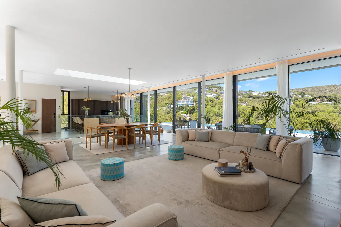 A bright, open-concept living space with beige sofas, a dining table, and floor-to-ceiling windows overlooking lush greenery.