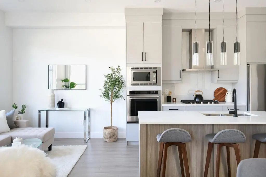 Bright, modern kitchen and living area with gray cabinets, white countertops, and wood-legged stools at the island. A gray sofa is visible.