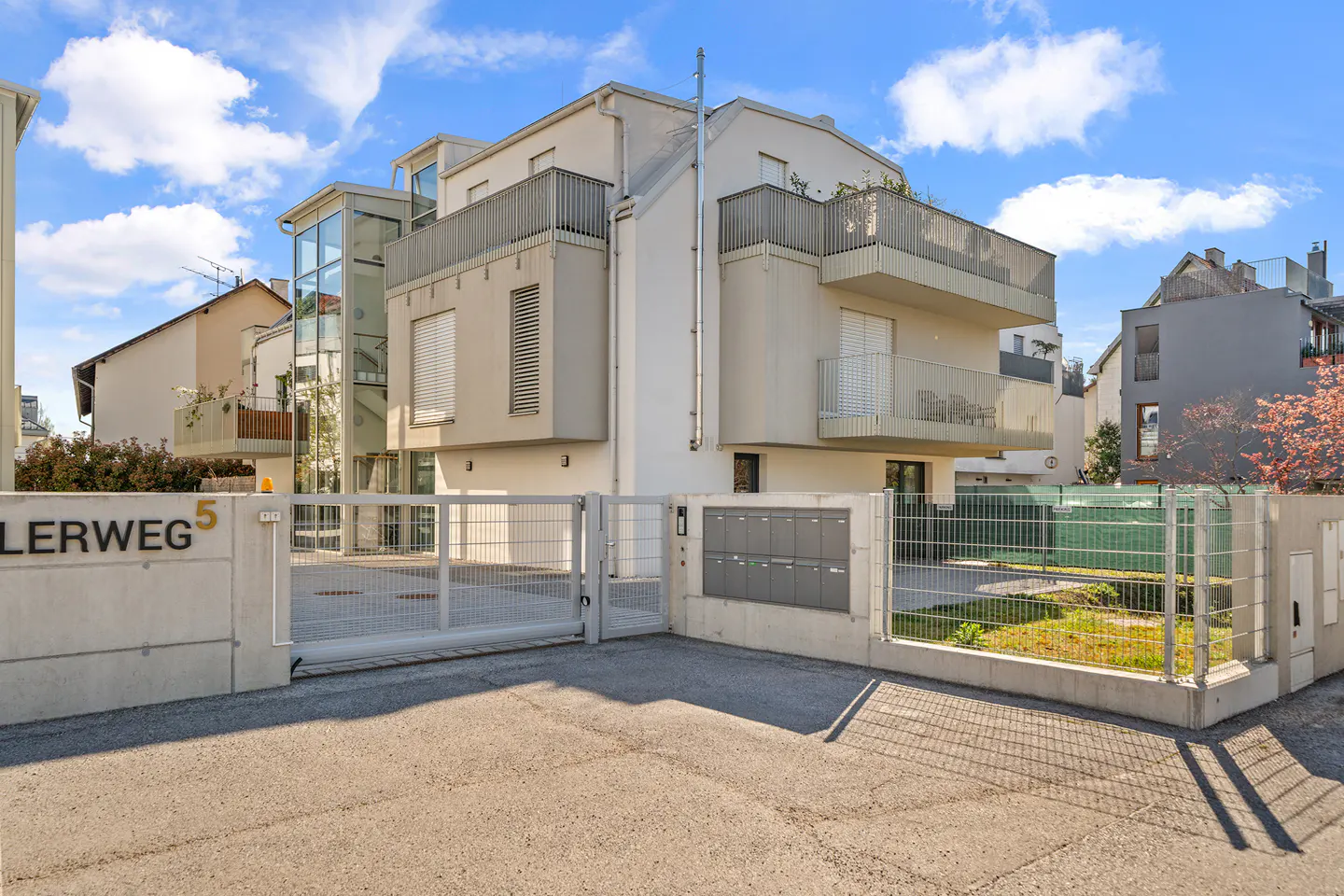 Exterior view of a modern, multi-story apartment building with balconies, a gated entrance, and a glass elevator shaft.