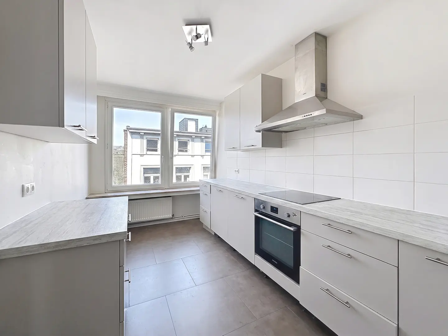 Bright kitchen with gray cabinets, white tile backsplash, and stainless steel range hood. A window overlooks a building.