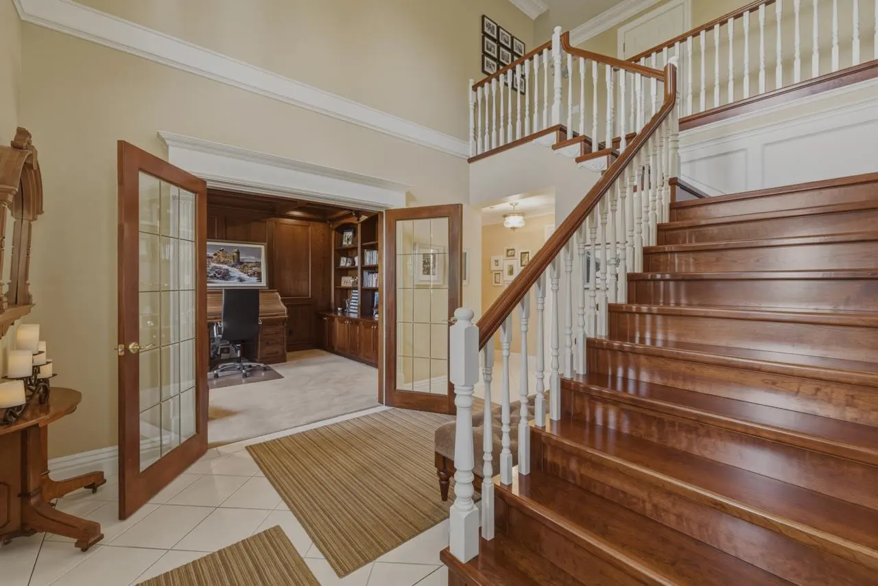 A foyer with a wooden staircase, white banister, and open glass doors to a wood-paneled office.