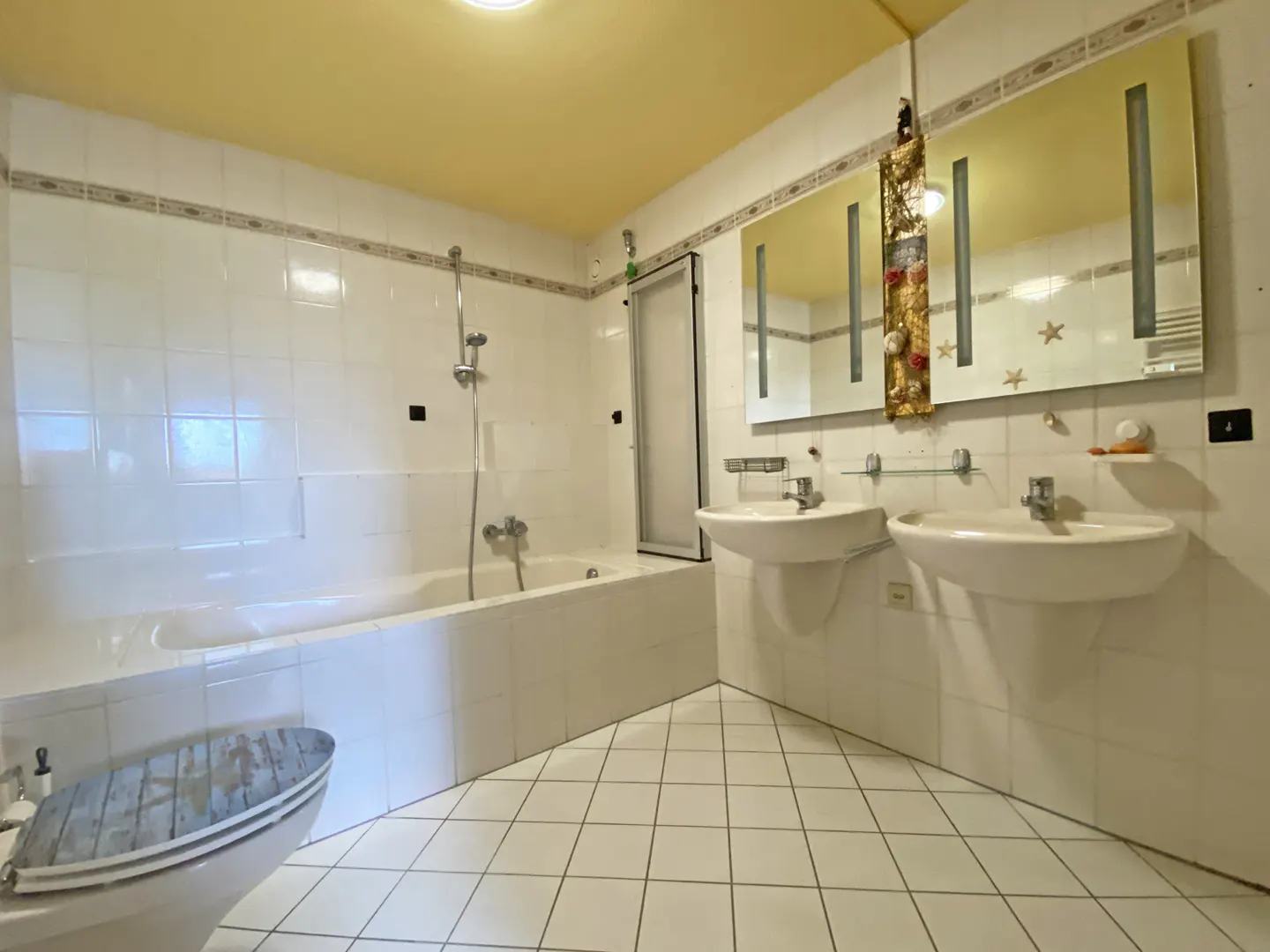 Bright bathroom with white tile, yellow ceiling, and two sinks. A bathtub with shower is on the left, and a toilet with a decorative lid is in the foreground.