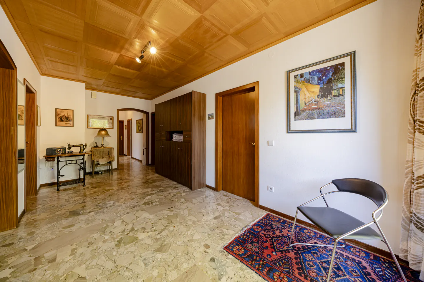 Hallway with a wooden ceiling, marble floor, and white walls. A chair sits on a red and blue rug. A painting hangs on the wall.