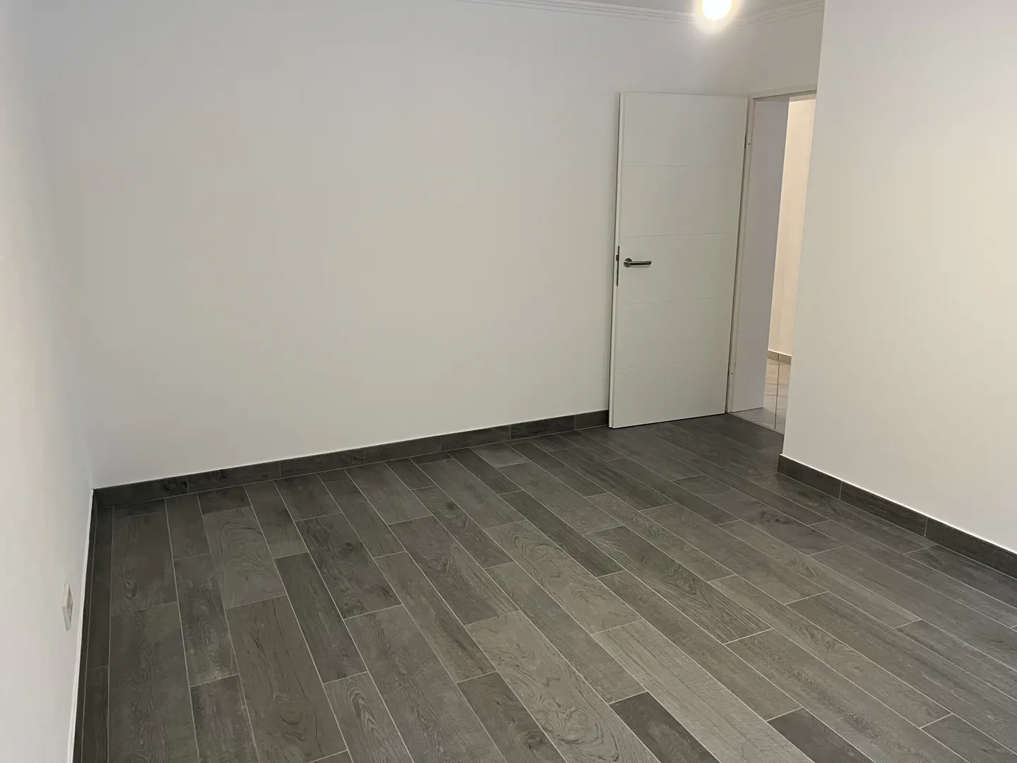 Empty room with white walls, gray wood-look tile floor, and an open white door leading to another room.