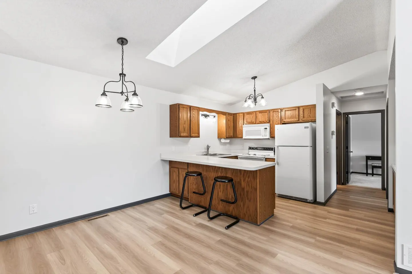 A bright kitchen with wood cabinets, white appliances, and a breakfast bar with two black stools. Wood-look flooring and white walls.