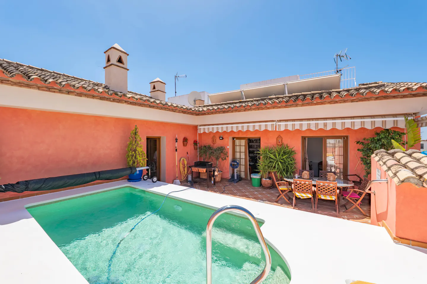 A sunny rooftop terrace with a small swimming pool, terracotta walls, and a dining area with chairs under an awning.