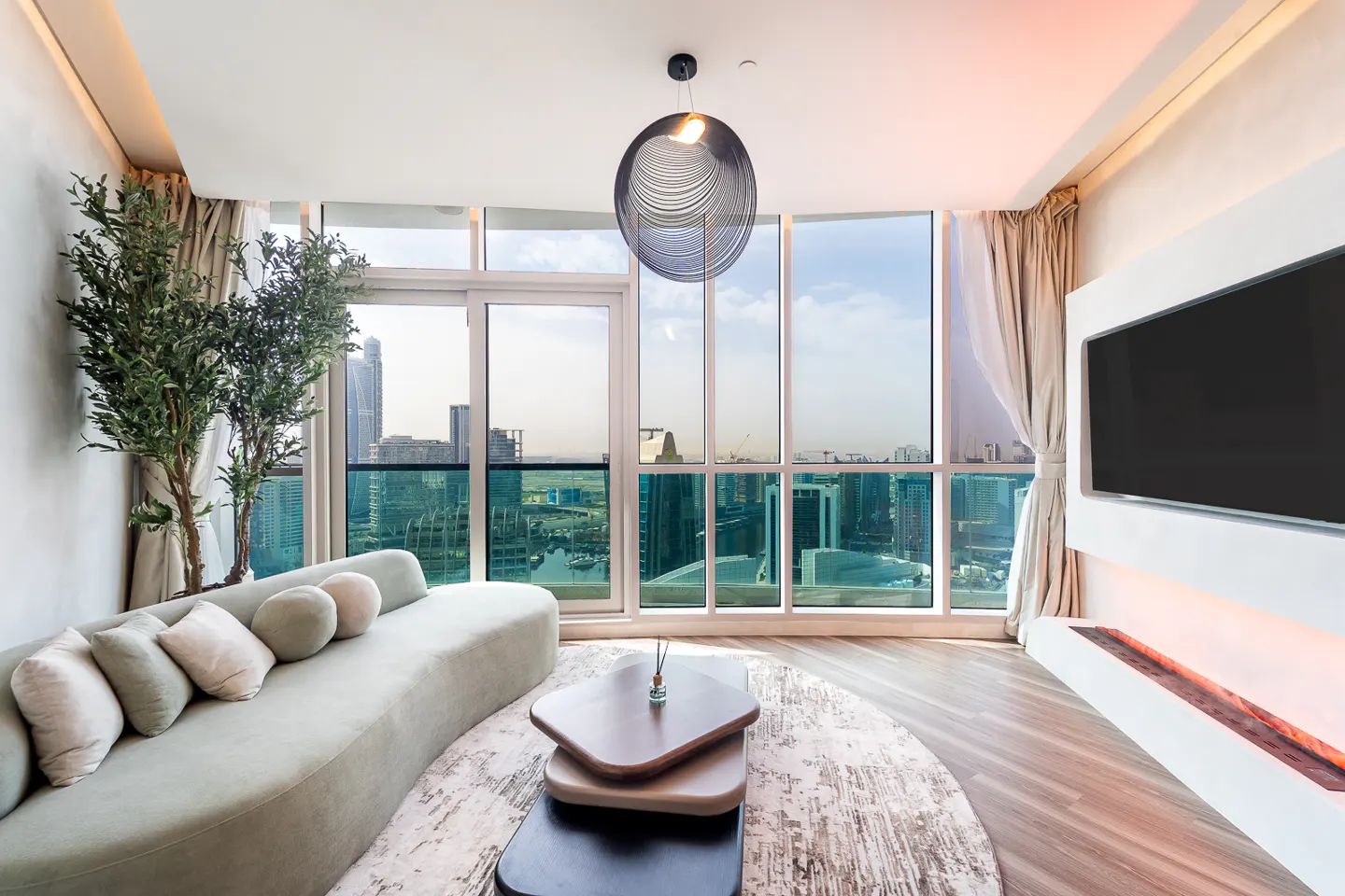 Bright living room with a curved sofa, layered coffee table, and city views from large windows. A modern light fixture hangs above.