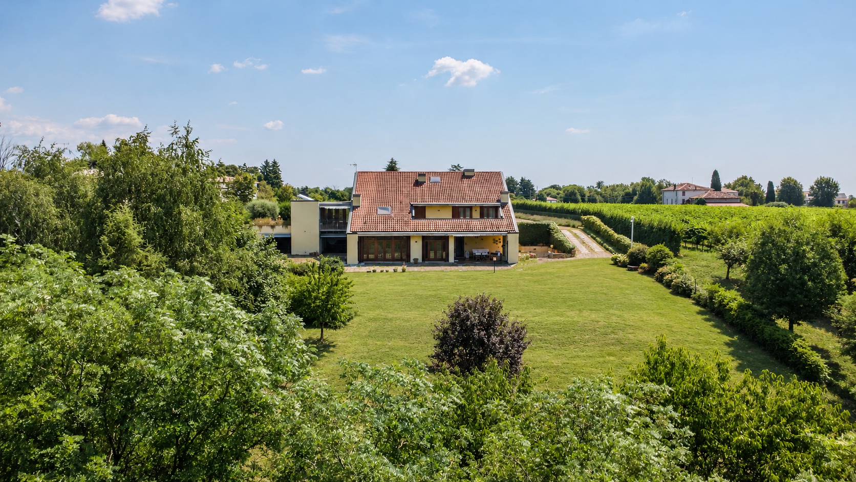 A two-story house with a red-tiled roof sits in a large, green lawn surrounded by trees and a vineyard under a blue sky. [AI-generated text]