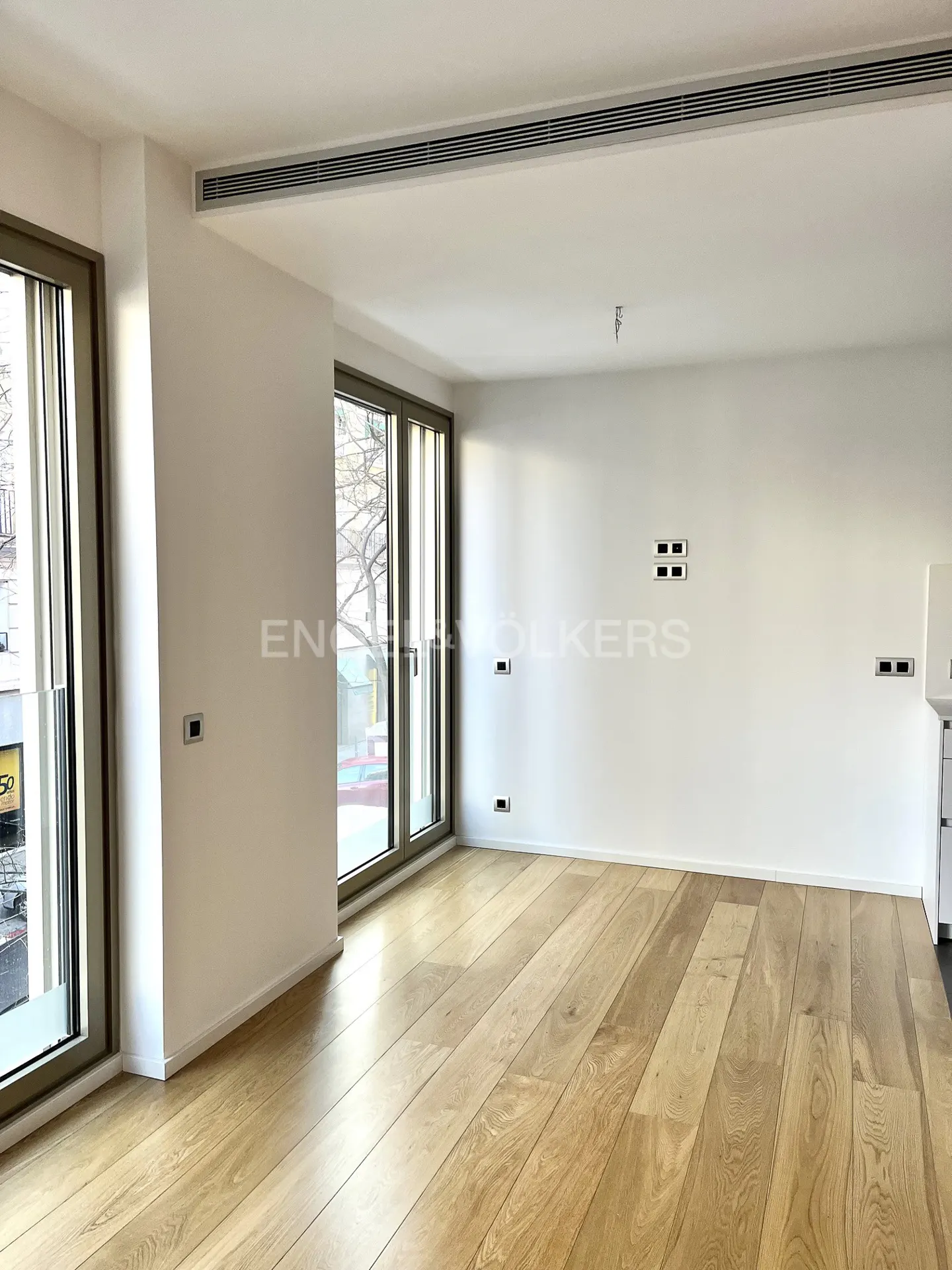 Bright, empty room with light wood floors, white walls, and large windows. Engel & Volkers logo visible on the wall.