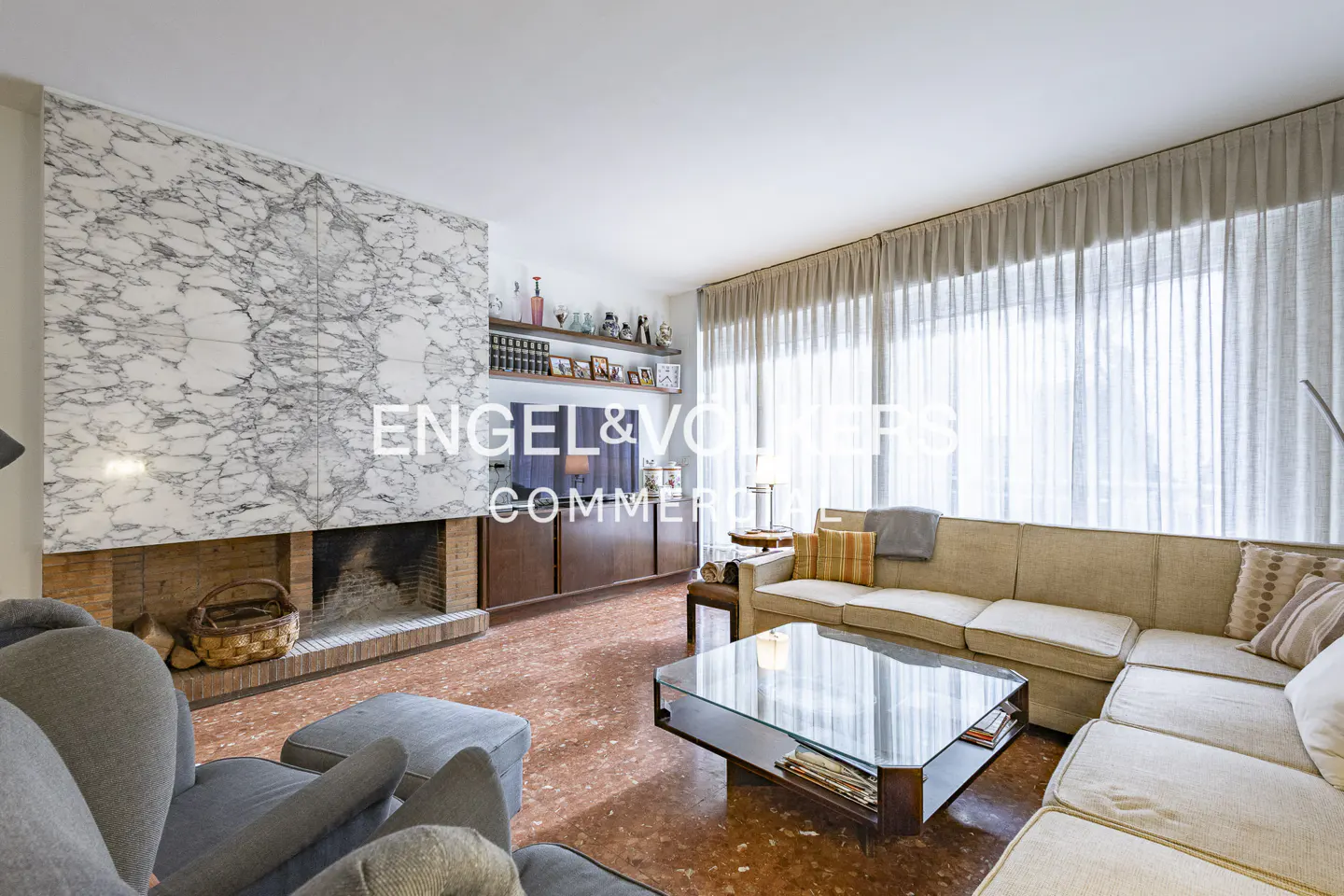 Living room with marble fireplace, beige sofa, glass coffee table, and red tiled floor. Large windows with sheer curtains.