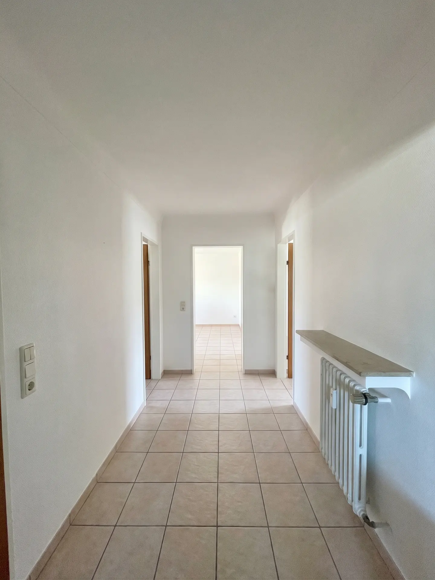 A bright hallway with white walls, beige tile floors, and open doorways leading to other rooms. A white radiator is visible on the right.