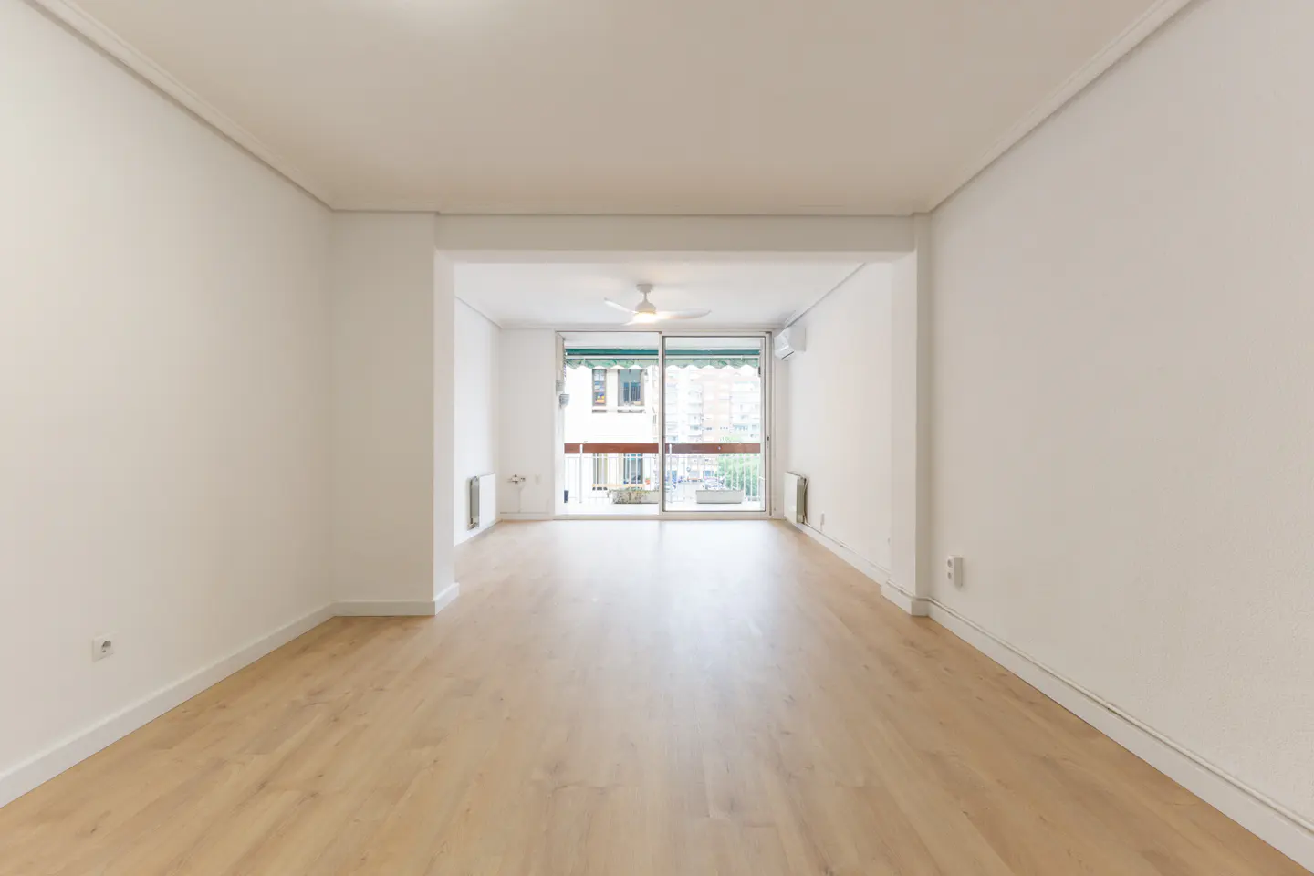 Empty, bright living room with white walls, light wood flooring, and a sliding glass door leading to a balcony.