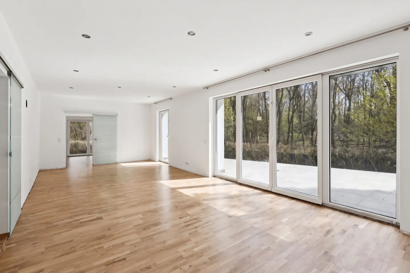 Bright, empty room with hardwood floors, white walls, and large sliding glass doors overlooking a wooded area.