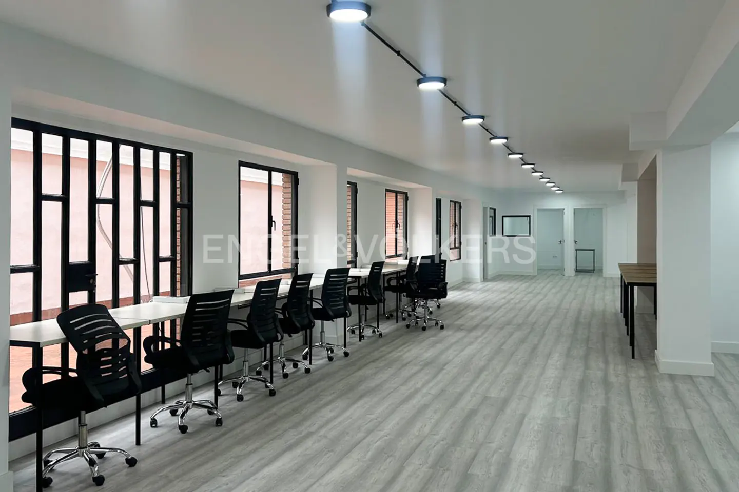 Bright, empty office space with white walls, gray wood floors, and black chairs at desks by windows. Overhead lighting.