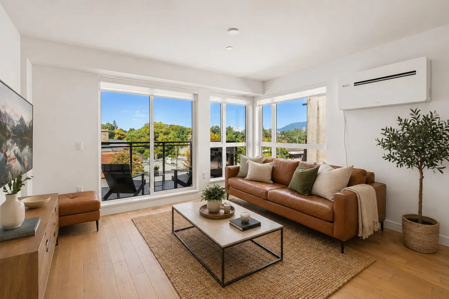 Bright living room with a brown leather sofa, jute rug, and large windows overlooking trees and a balcony.
