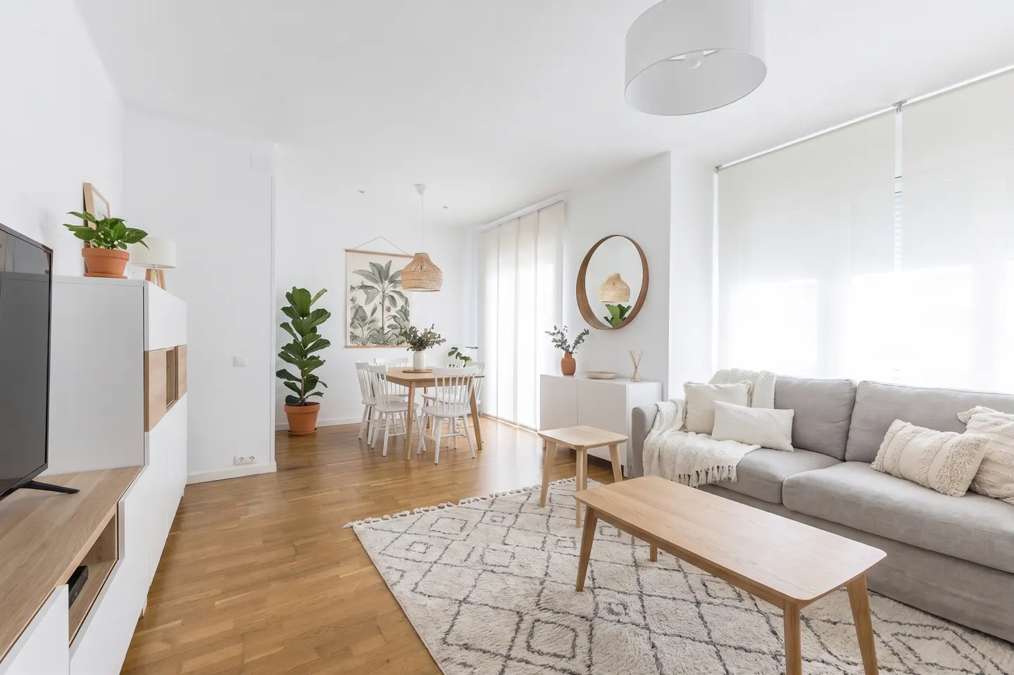 Bright living room with a gray sofa, wooden coffee tables, and a dining area with white chairs. A patterned rug lies on the wooden floor.