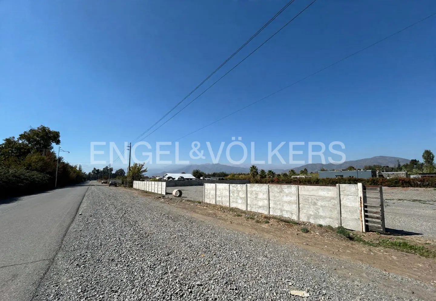 A vacant lot with a concrete wall and a gravel road under a clear blue sky. Mountains are visible in the background.