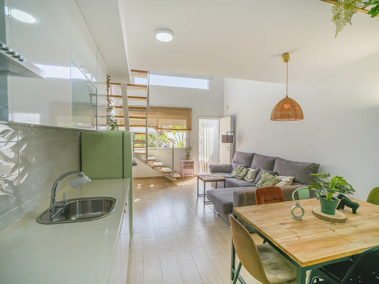 Bright, open-concept living space with kitchen, dining table, and gray sofa. A wooden staircase leads to an upper level. Natural light fills the room.