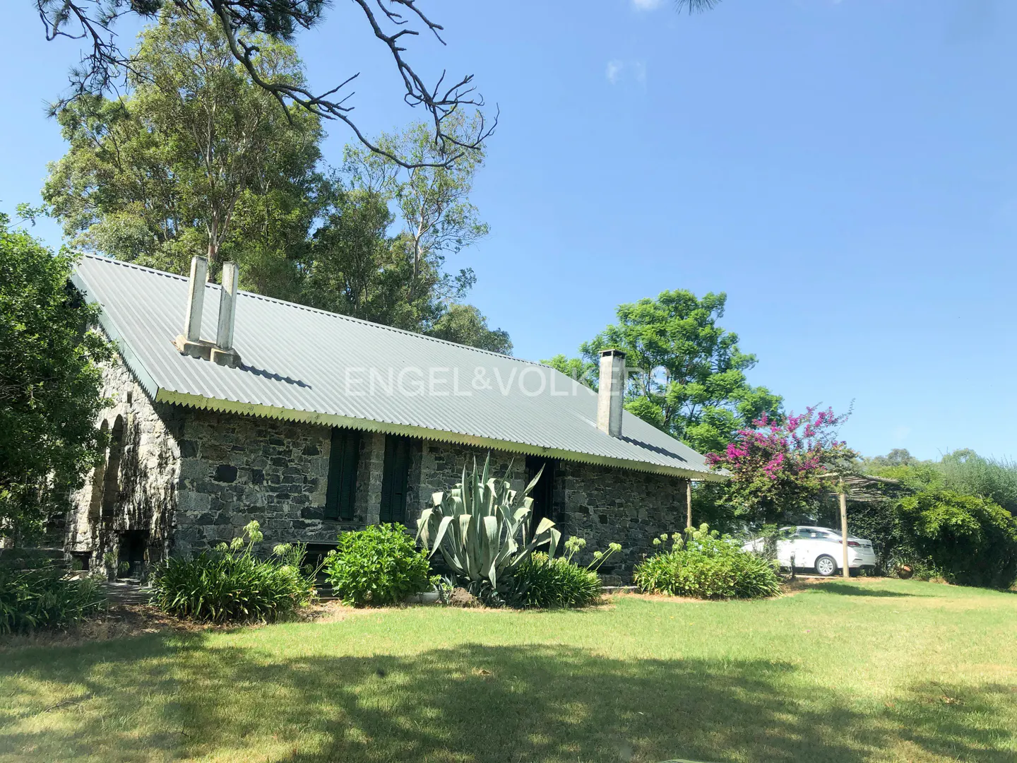 Stone house with a metal roof, green shutters, and two chimneys, surrounded by green grass, trees, and a blue sky.