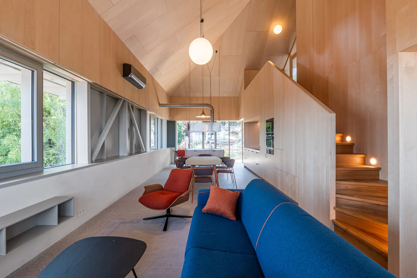 Bright, modern interior with wood walls, blue sofa, and orange chair. Stairs lead up to a second level. Large windows offer natural light.