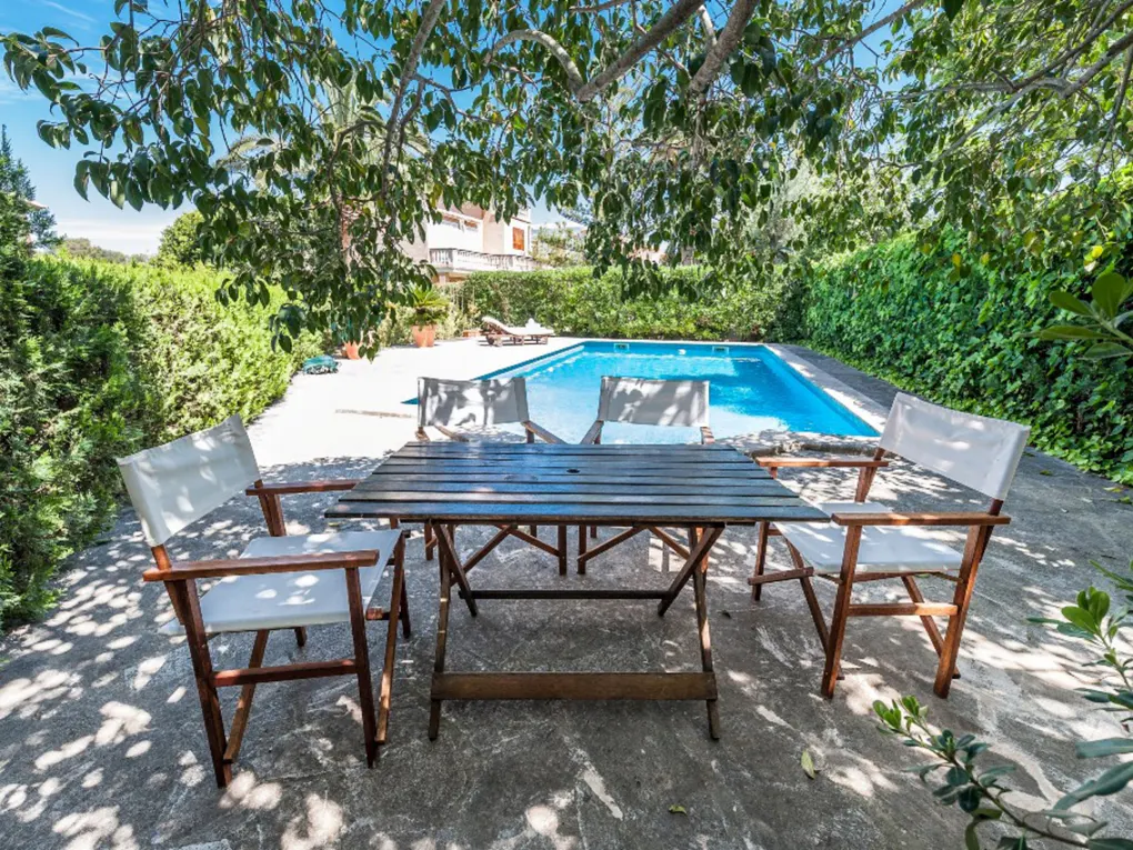 Wooden table and white chairs on a stone patio overlooking a bright blue swimming pool surrounded by lush green hedges.