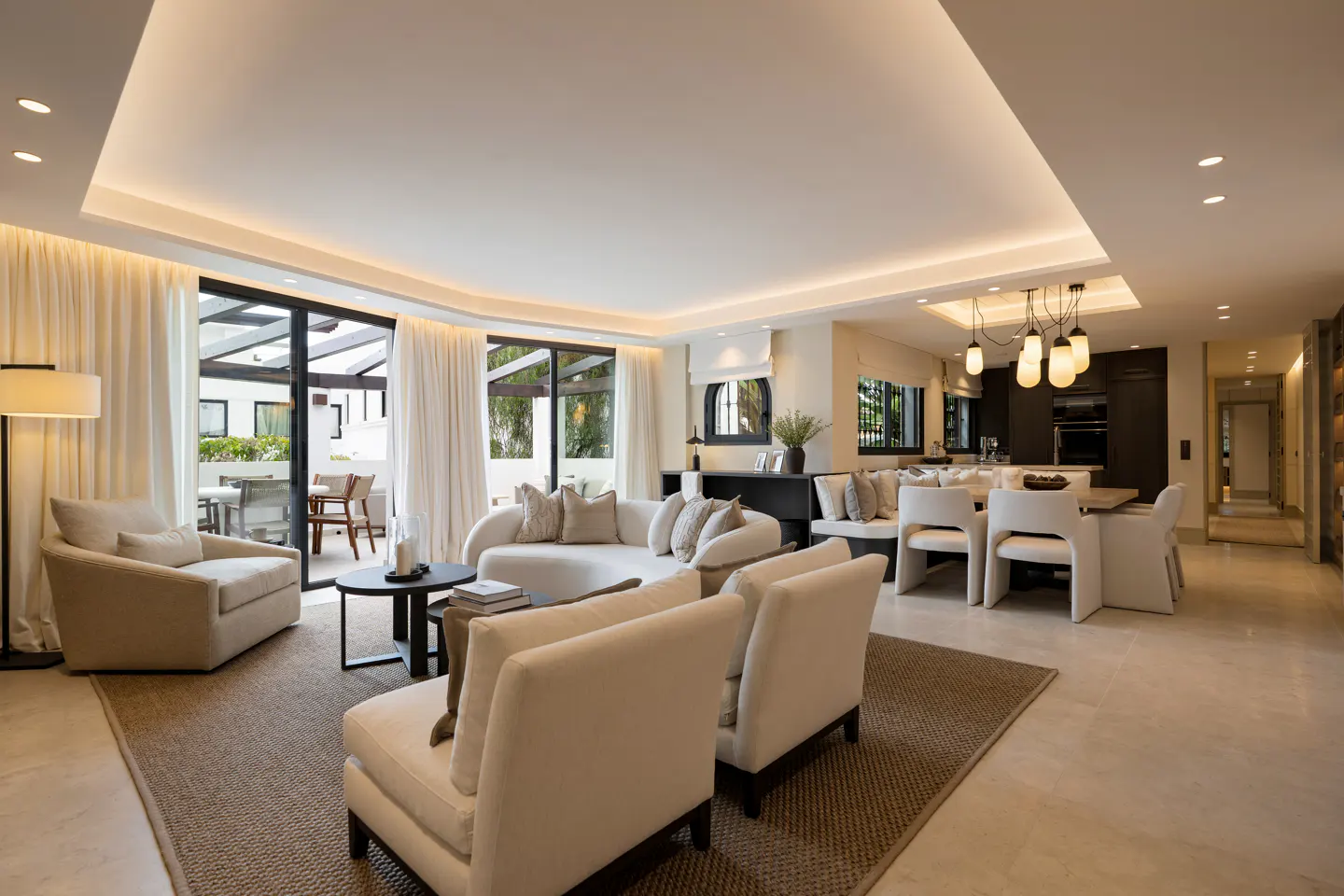 Bright, open-plan living room with neutral tones. Cream sofas and chairs sit on a brown rug, with a dining area and kitchen visible in the background.