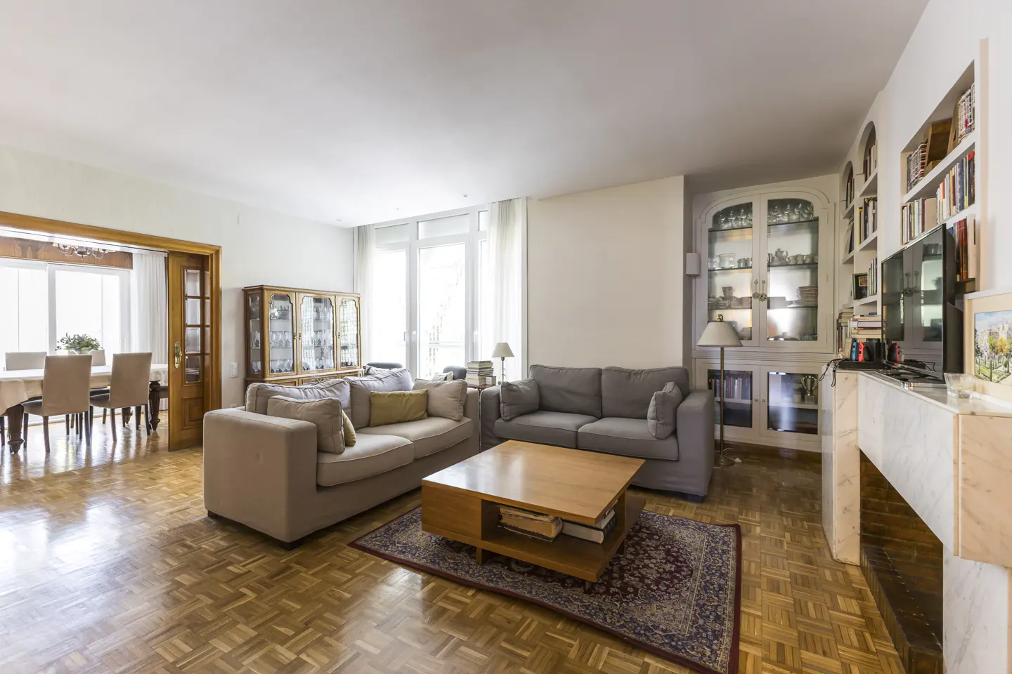 Bright living room with parquet floors, two grey sofas, a wooden coffee table, and built-in shelving with a TV.