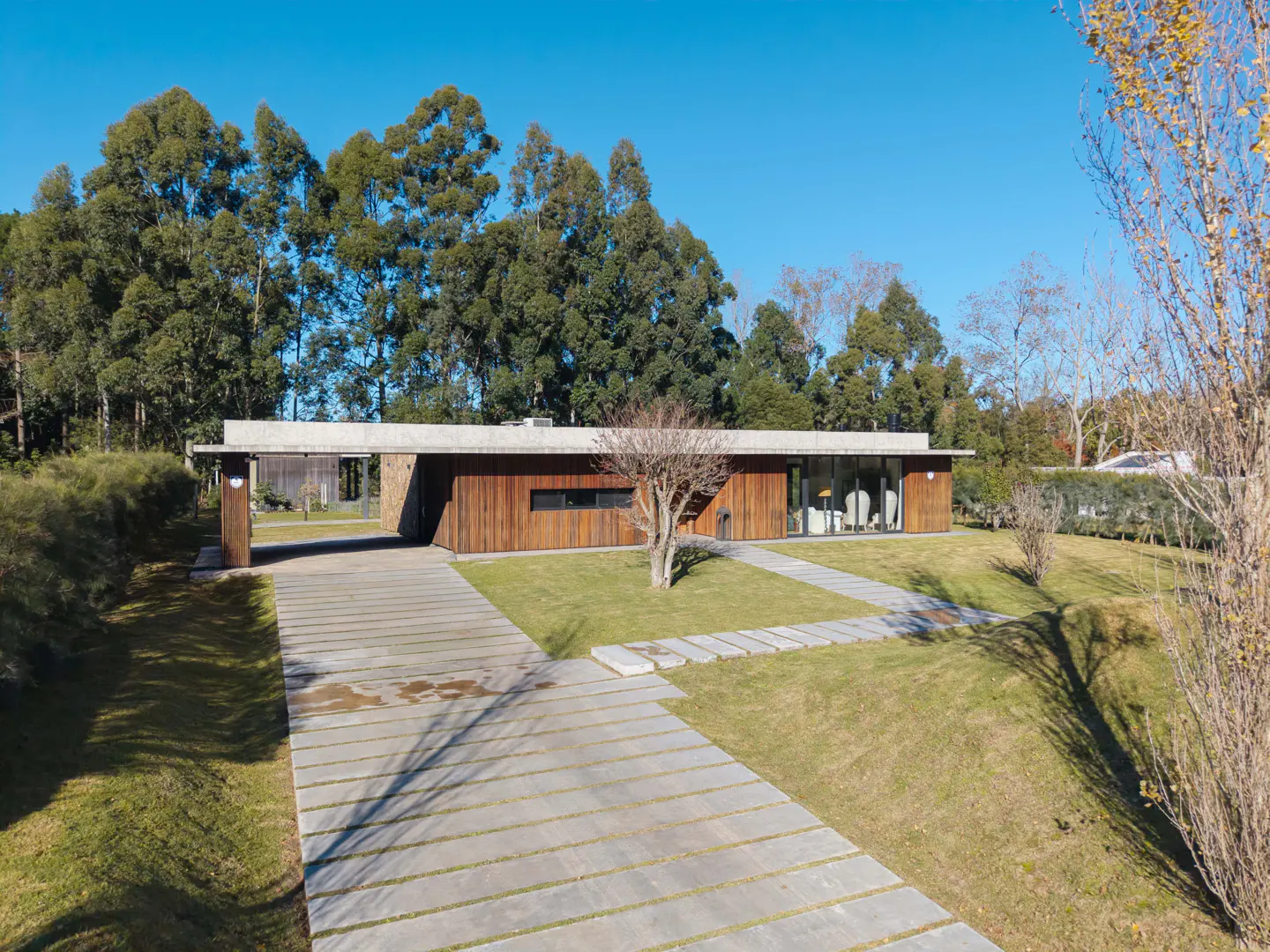 Modern single-story home with wood siding, concrete roof, and a stone driveway set in a lush, tree-lined landscape.